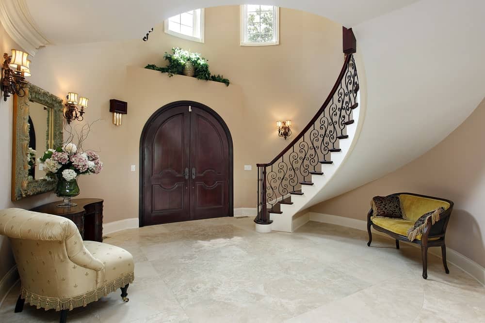 这个意大利别墅风格的门厅里有一个装饰性的栏杆，一个毛绒丝绸沙发和一个大花瓶，突出了苍白的大理石地板和柔和的米黄色墙壁。