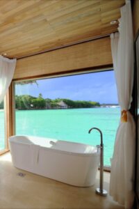 热带主浴室设有一个独立的浴缸，前面有一个大窗户，可以看到海洋的景色。这个空间还包括木质天花板、瓷砖地板和带水槽的梳妆台。