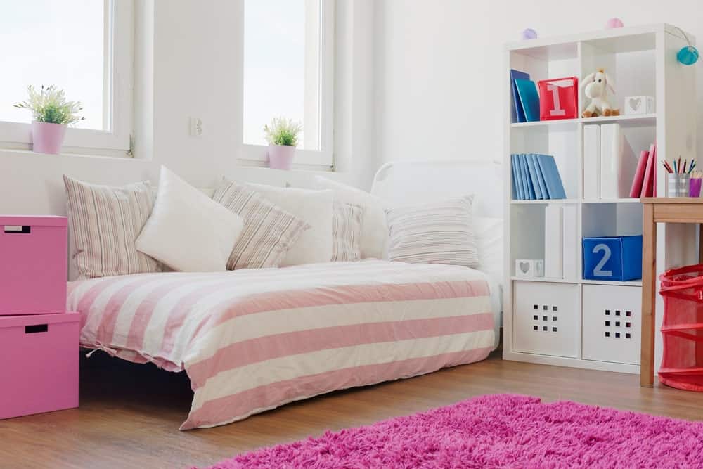 这个女孩的卧室以白色的墙壁和粉红色为主调。床边有一个高高的搁板。一边是书桌的位置。