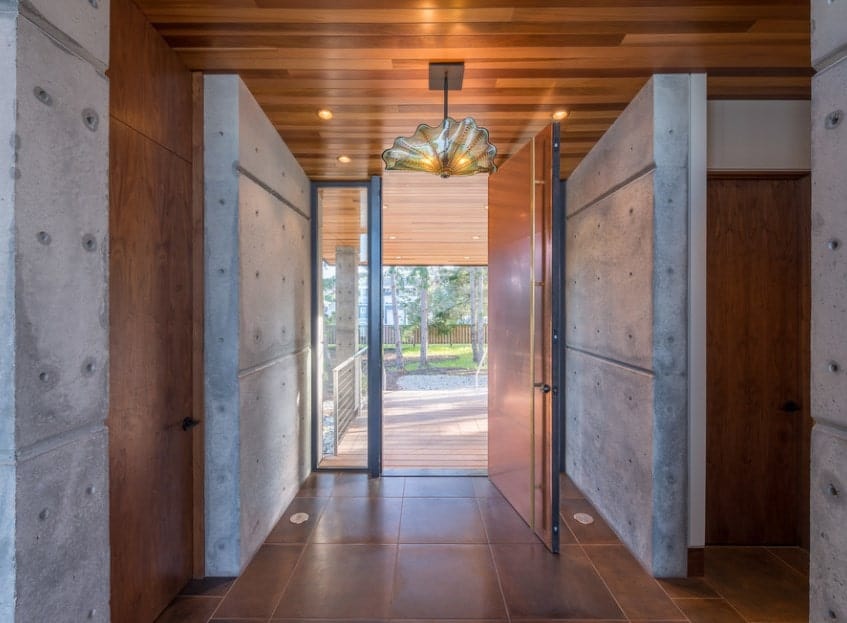 这间现代风格的门厅铺着土褐色地砖，与深棕色的主门、墙板和木质天花板相匹配。灰色的混凝土墙与之相得益彰，从敞开的门和玻璃侧灯射进来的自然光使其更加明亮。