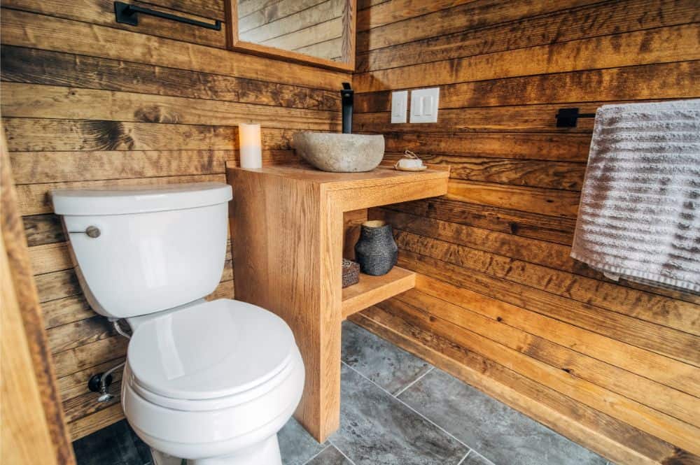 一间小房子里的乡村浴室，有破旧的木墙，定制的木质梳妆台和混凝土容器盆，旁边是白色的厕所。