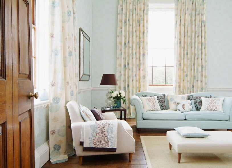 美丽破旧别致的浅蓝色墙壁与两扇明亮的窗户上令人难以置信的彩色米色窗帘相辅相成。这也补充了浅蓝色沙发，搭配米色软垫扶手椅和软垫奥斯曼咖啡桌。