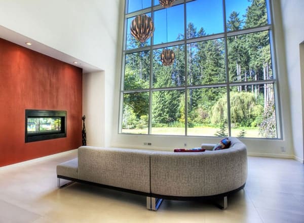 这是一间宽敞通风的客厅，里面有一张灰色弯曲的大组合沙发，一面巨大的玻璃墙，土红色墙壁上有一座现代化的壁炉。