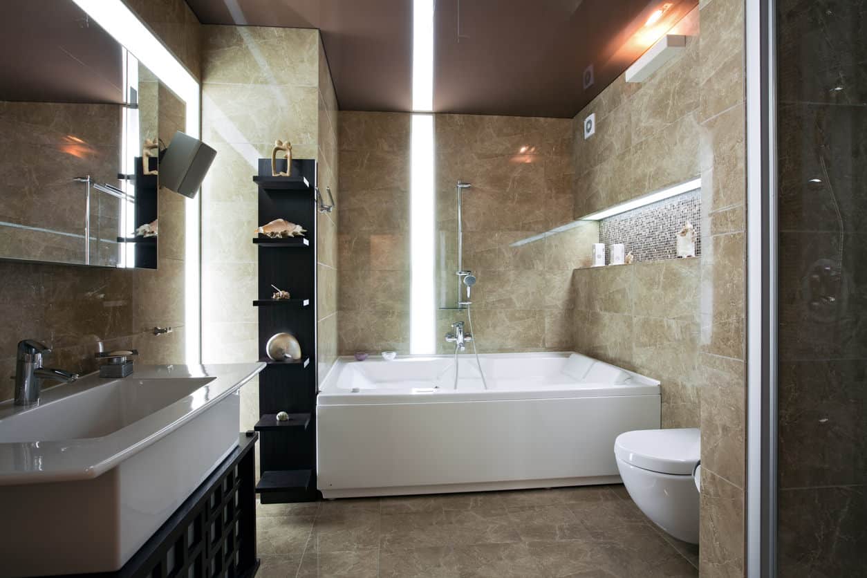 这是一个男性化的浴室设计，有一个巨大的白色槽水槽垂直于凹形白色浴缸。浴缸上方有一个凹槽架，用来放洗发水、肥皂和其他产品。这里一个突出的设计特点是灯光。
