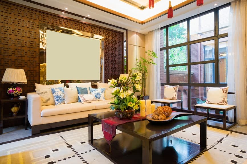 白色软垫沙发后面的墙上有东方图案，装饰着金色框架的空白帆布，与带有装饰和盆栽的大型黑色木制咖啡桌下面的大型图案区域地毯相匹配。