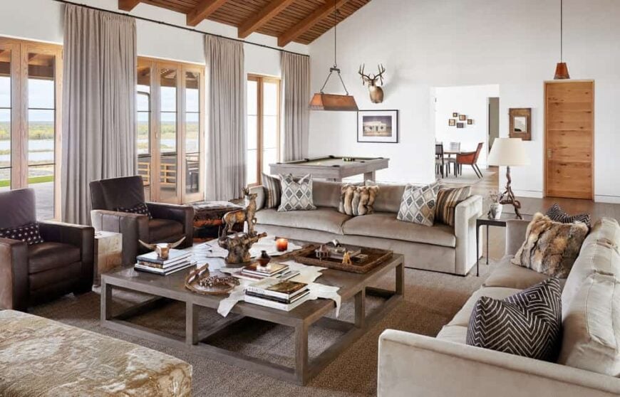 这是一间乡村风格的客厅，米色软垫沙发和扶手椅环绕着一张大木制咖啡桌，上面有高高的木制横梁天花板。