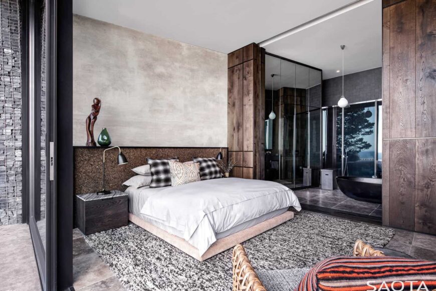 当代风格的主卧室以一张低矮的床为特色，床放在灰色地毯上，上面是石头瓷砖地板。它有一个深色的木床头柜和台灯。