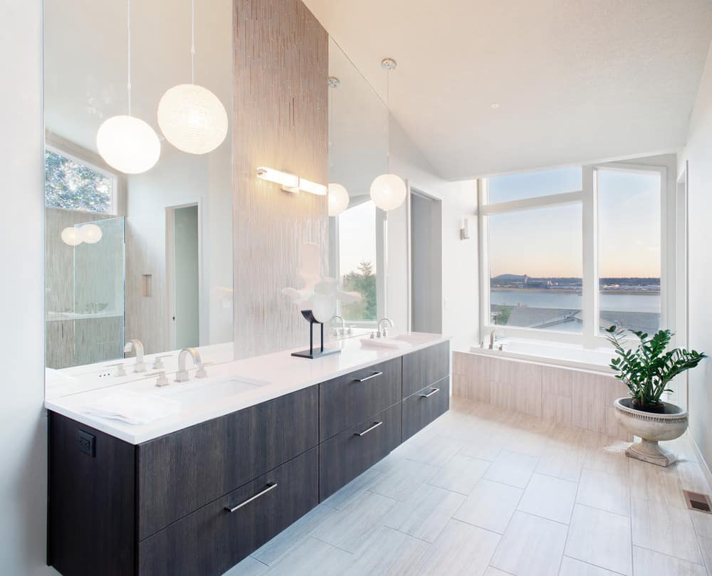 光线充足的现代主浴室，尽头有一个凹形浴缸，在一个可以看到海洋的大窗户下。我喜欢这个主浴室，因为它的简单和漂浮的深色木梳妆台与剩下的白色和非常柔和的颜色之间的鲜明对比。