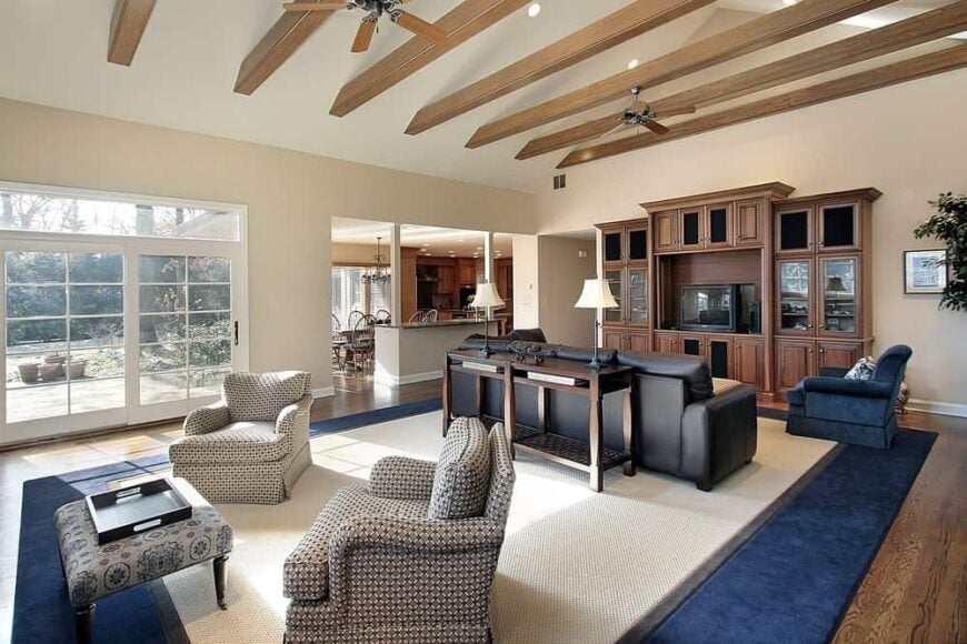 宽敞的工匠客厅设有拱形天花板和裸露的木梁，以及带有天鹅绒蓝色边框的奶油色地毯，与硬木地板上的扶手椅相匹配。