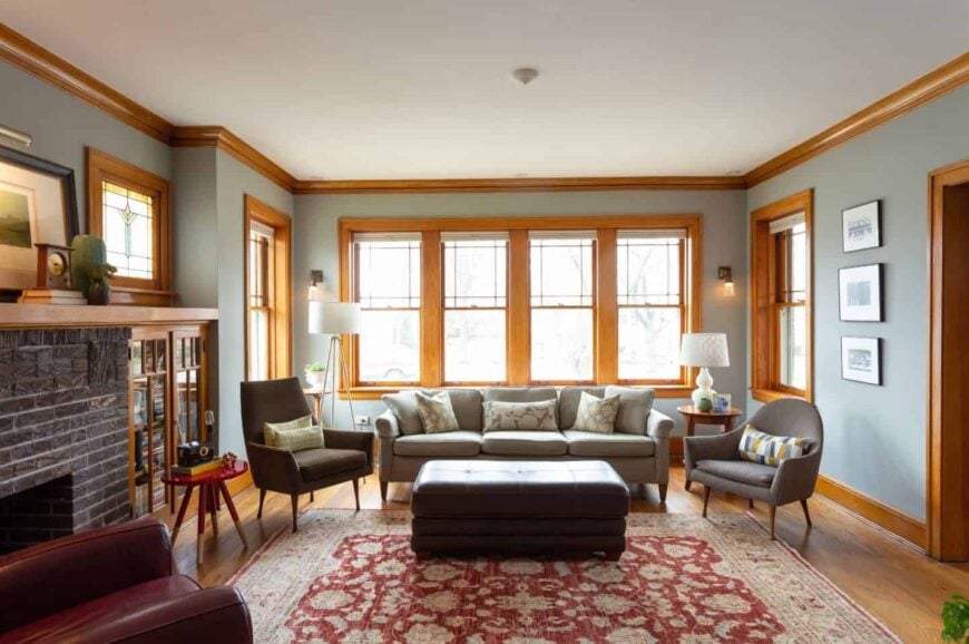 灰色的客厅以木制的冠饰和窗框为特色。房间里有灰色的组合式沙发和扶手椅，中间有一张搁在红色复古地毯上的软脚凳。
