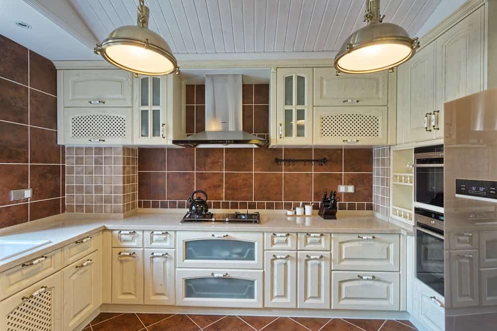 这是一间经典的厨房，墙壁和地板都是棕色的瓷砖，白色拱形天花板上挂着两盏巨大的吊灯，还有白色的橱柜和不锈钢用具。