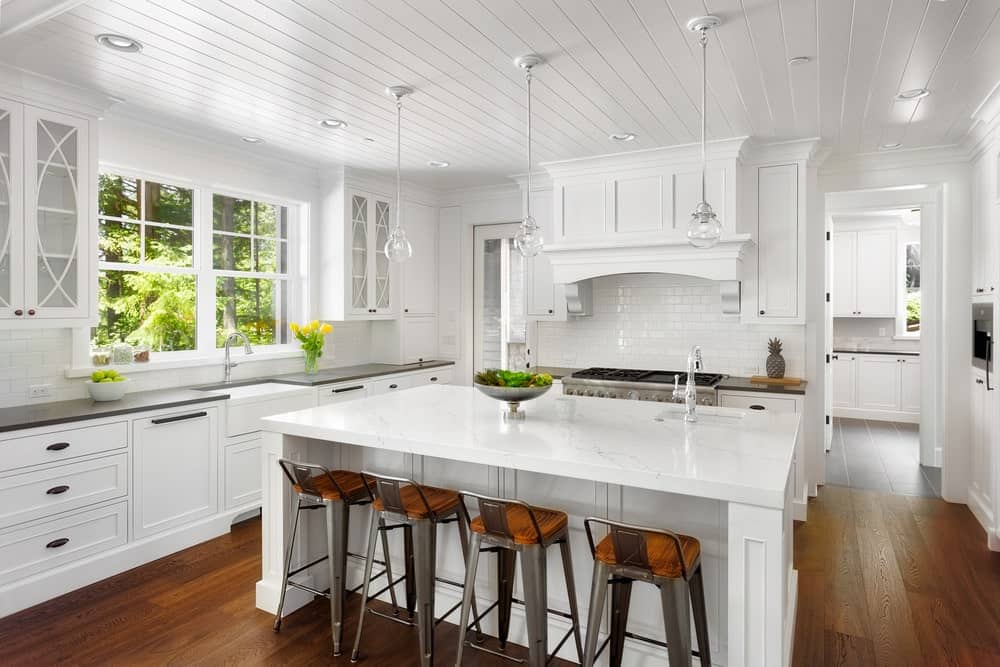新鲜的厨房有白色的橱柜，砖砌的后挡板，硬木地板，白色的岛式桌子和椅子，白色的天花板和悬挂的玻璃吊灯。