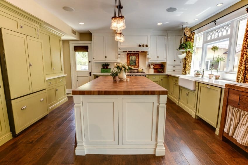 简单的厨房，硬木地板，白色和橄榄绿的橱柜，一个木制的岛式桌子和一盏玻璃吊灯。