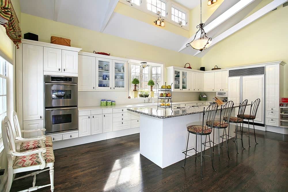 明亮的厨房有黄色的墙壁，硬木地板，白色的橱柜，长方形的岛式桌子和椅子，复古的吊灯。
