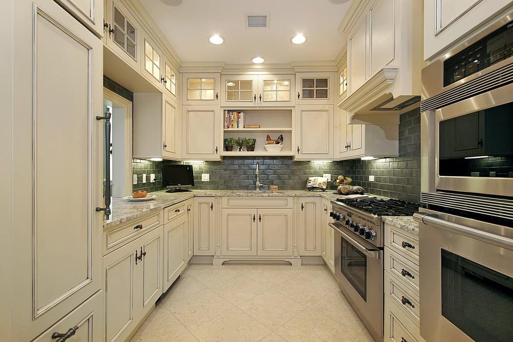 厨房里有黑色的砖挡板，灰色的大理石台面，白色的橱柜和架子，瓷砖地板，嵌入式照明和不锈钢电器。
