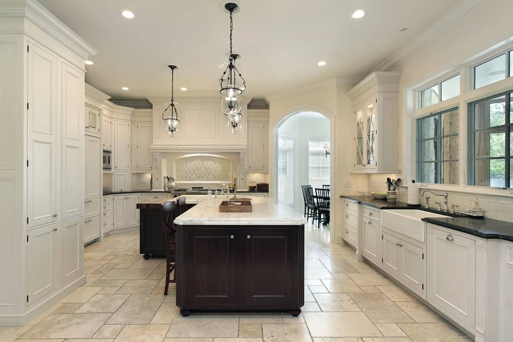 白色干净的厨房里有木制橱柜，一张带白色大理石台面的岛式餐桌和深棕色的橱柜，铺着瓷砖的地板和一盏复古的吊灯。