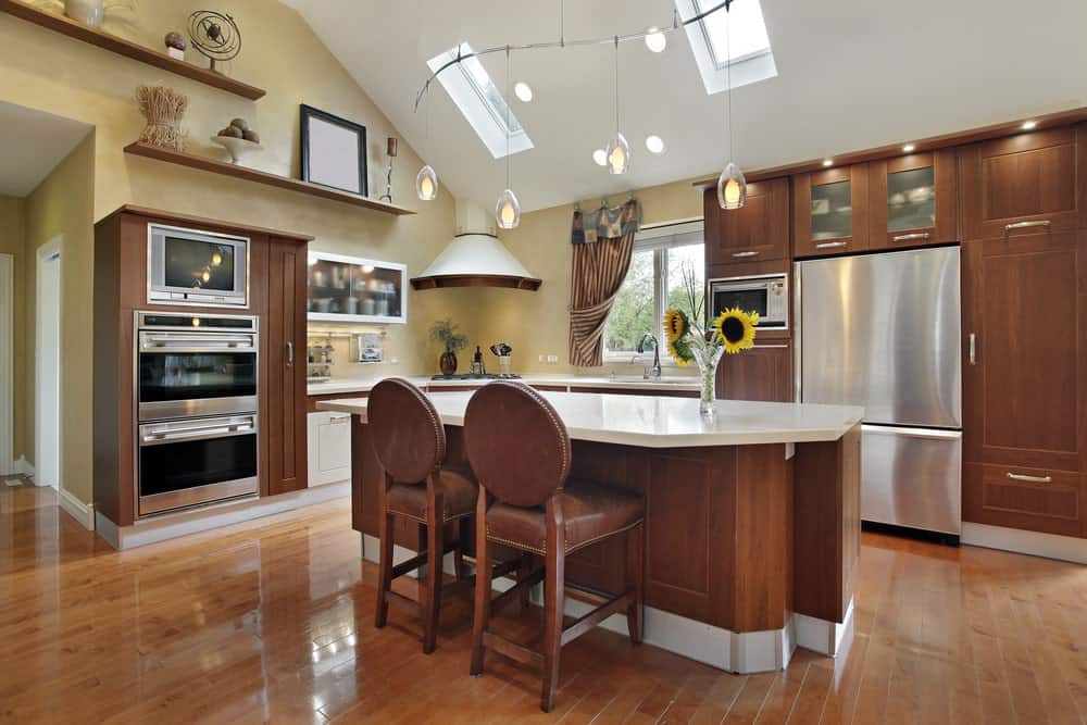 舒适的厨房，拱形天花板上挂着吊灯，一张岛式桌子，两把软垫椅子，棕色木橱柜和架子。