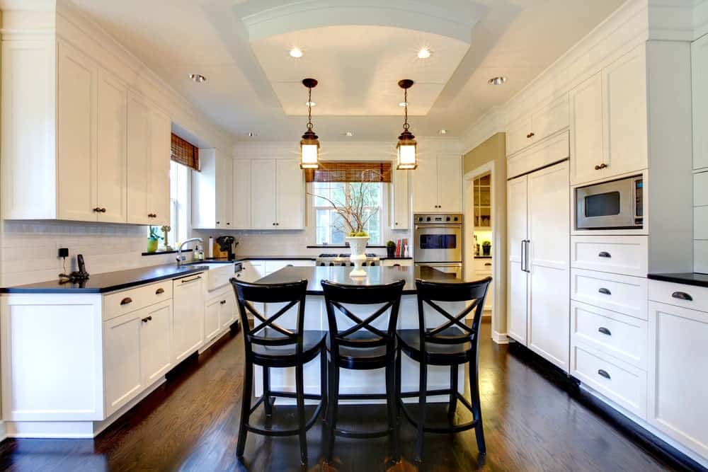 光滑的厨房有白色木橱柜，光滑的白色砖后挡板，优雅的吊灯，硬木地板和一个带三把木椅的早餐岛。
