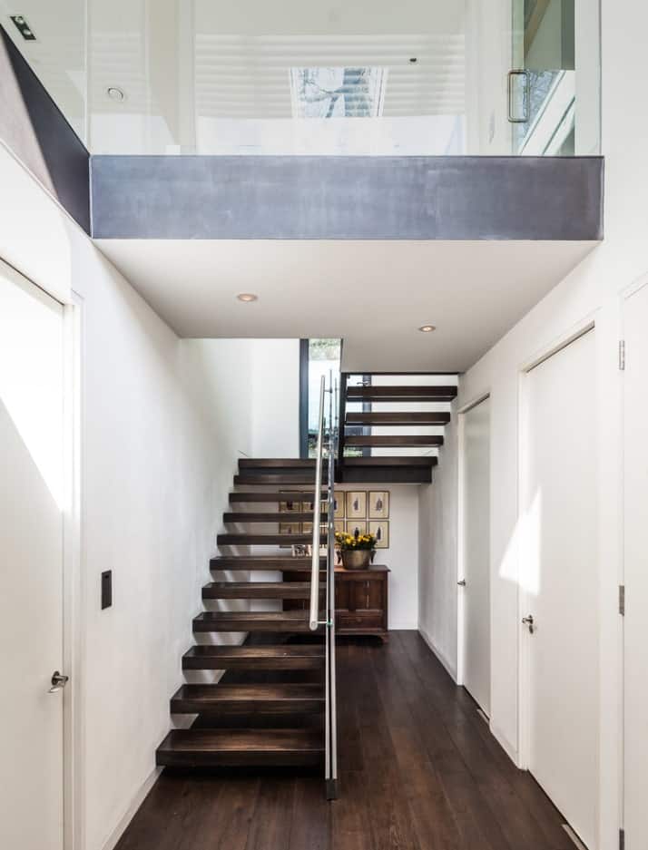 现代楼梯，深色木踏板，开放式立管和钢扶手沿着白墙。为了节省空间，它下面有一个木柜子。