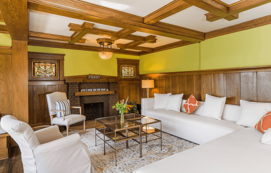 一个舒适的工匠客厅，黄色的墙壁上的木板墙补充了格子木天花板梁。它的特点是白色的l形沙发以及玻璃中心桌和白色扶手椅。