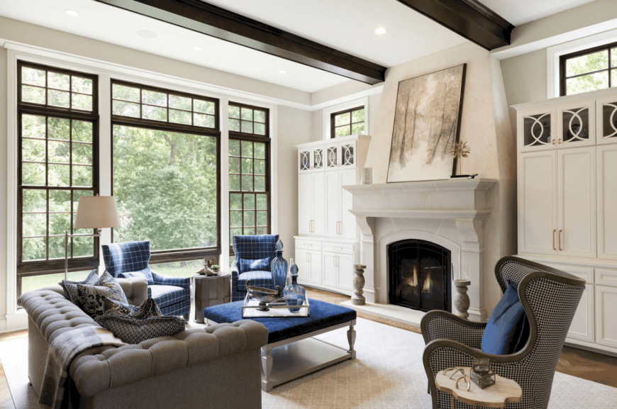 白色工匠风格的客厅由灰色簇绒沙发和蓝色方格扶手椅突出，凸显了天鹅绒蓝色的中心桌子。雅致的白色橱柜之间有一座现代化的壁炉。