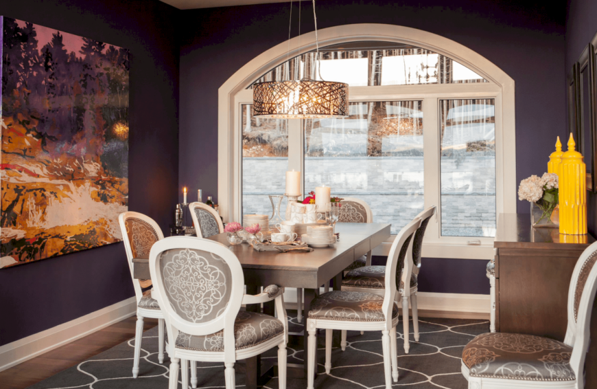 一个紫色的餐厅与流行的黄色口音，时尚的灰色软垫椅子搭配灰色木桌，和一个独特的吊灯。