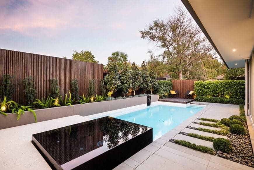 极简主义风格的游泳池配有黑色马赛克瓷砖，以木墙为特色，在木质和修剪过的灌木墙上设有混凝土种植箱。