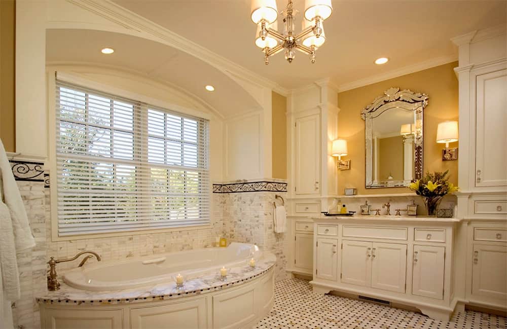 黄色浴室带有微妙的金色暗示，窗户下的角落浴缸毗邻带金色镜子的白色梳妆台。它由壁式烛台和一盏可爱的枝形吊灯照亮。