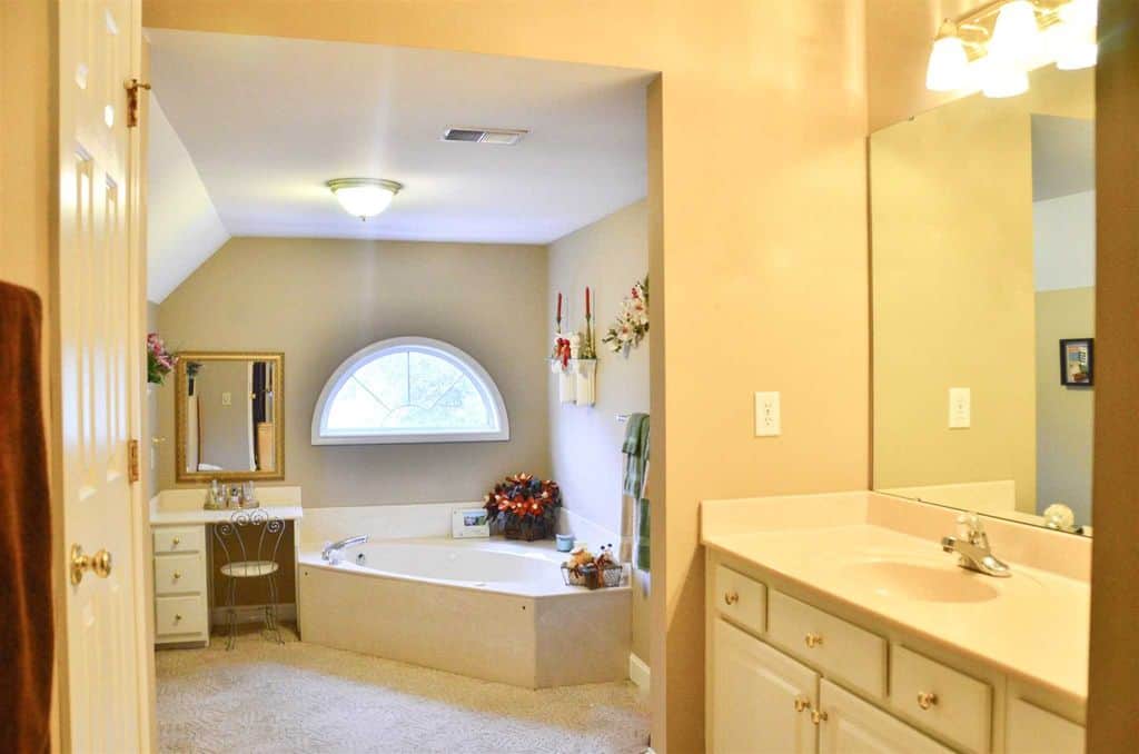 温暖，黄色的浴室设有一个半圆形窗户下的角落浴缸，坐在地毯地板上。旁边是一张小梳妆台，配有金镜子和金属椅子。