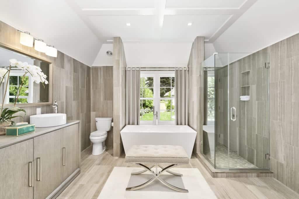 带有金属底座的簇绒软脚凳，旁边是白色框窗下的独立浴缸，是这个舒适浴室的焦点。