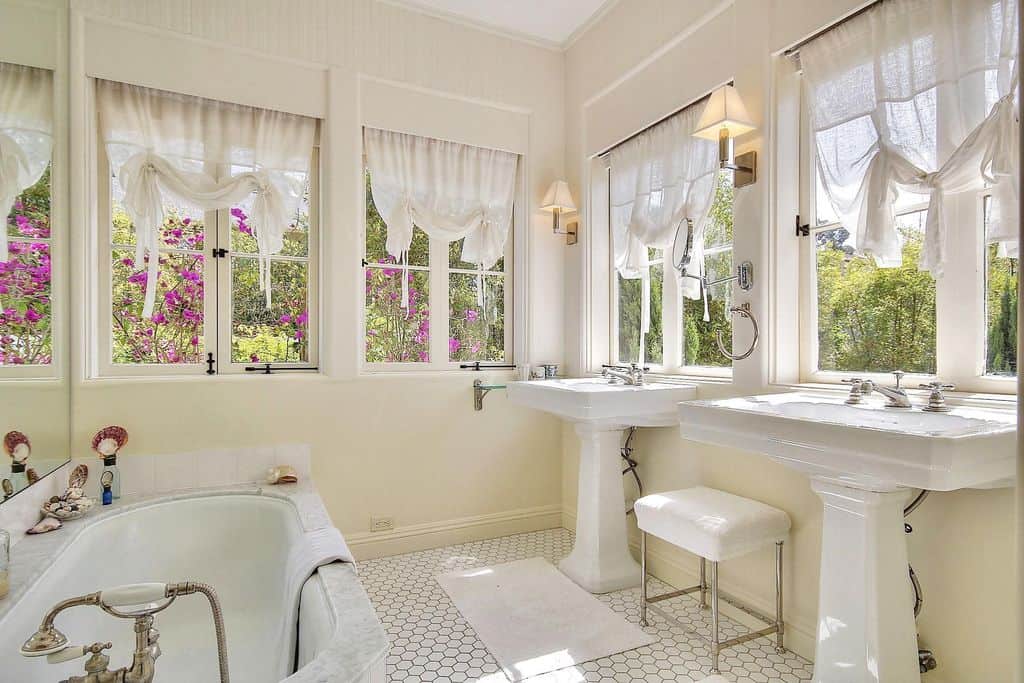 明亮通风的浴室设有一对底座式洗衣箱，中间有白色凳子，面向大理石浴缸。它被框窗包围，为这个中型主浴室增添了一种新鲜感。