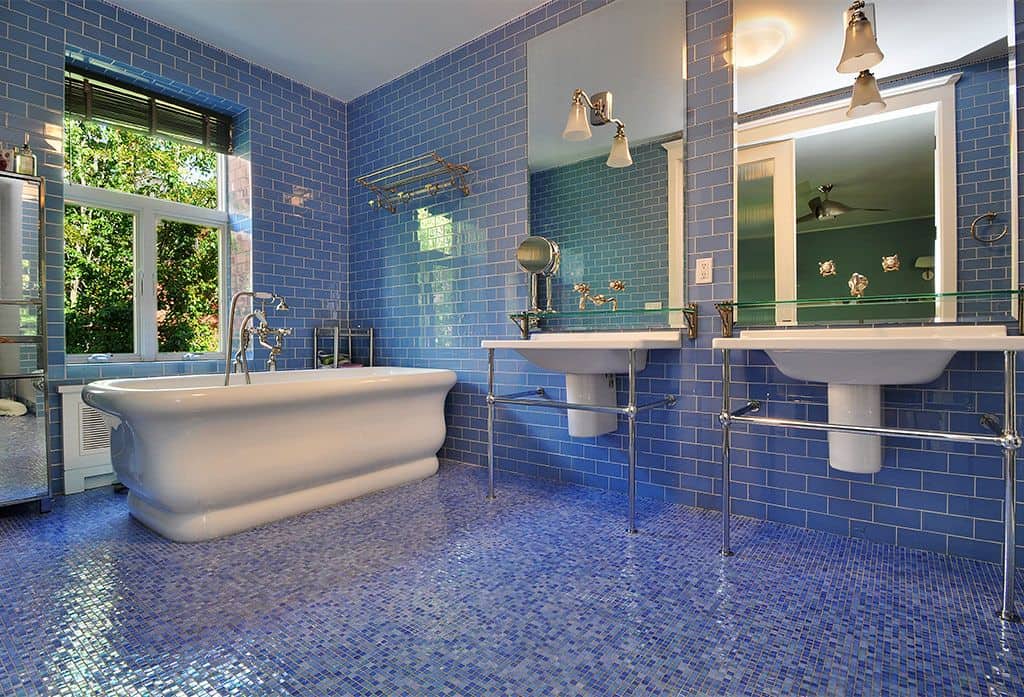 凉爽的浴室，蓝色地铁瓷砖墙面搭配蓝色马赛克瓷砖地板，一个独立浴缸和一对浴室水槽，由安装在无框镜子上的壁灯照明。