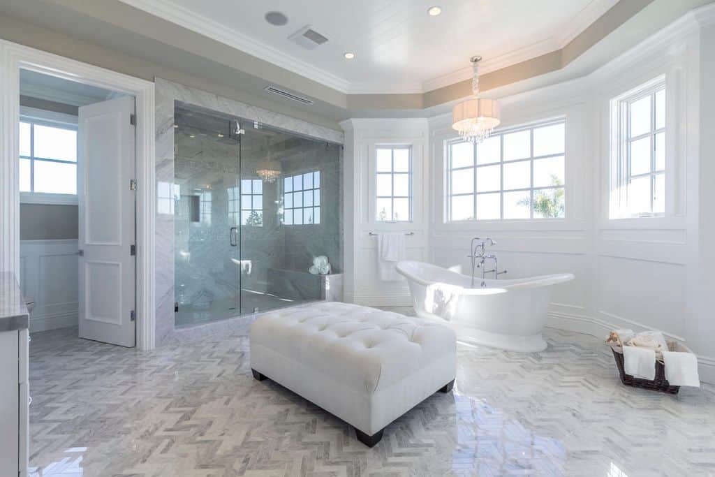 豪华的白色浴室设有一个独立的浴缸，旁边是白色的簇绒搁脚凳，上面有光滑的人字纹地板。它由一盏悬挂在凹形天花板上的花式吊灯照亮。
