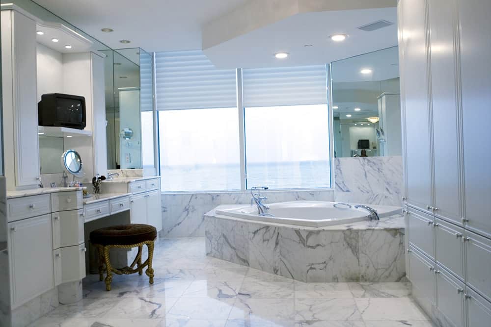 灰色大理石覆盖了这个光滑的中型浴室的地板、墙壁和浴缸。它有一个带有棕色软垫的凳子的梳妆台，面对着一个巨大的灰色柜子，通过窗户可以看到海滩的壮丽景色。