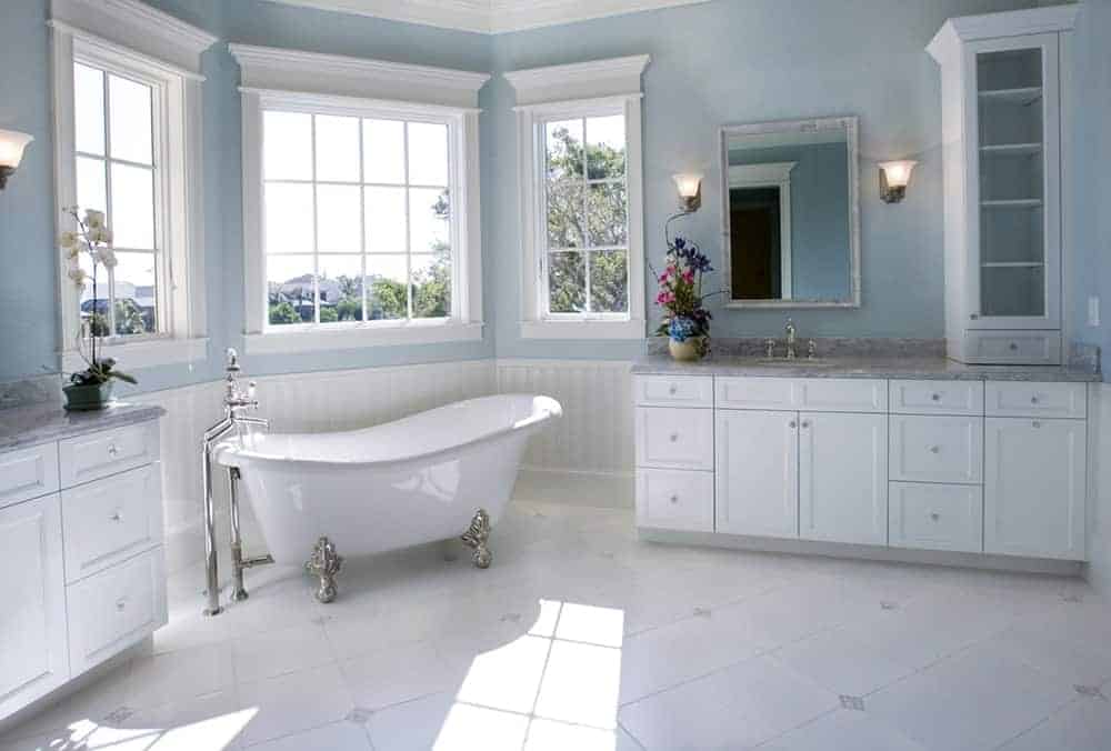 可爱的白色和蓝色浴室，白色木脸盆架中间有一个独立的爪足浴缸，上面有灰色大理石。