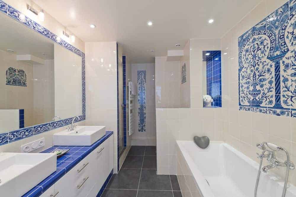白色瓷砖浴室强调蓝色平原和图案瓷砖在墙壁上，台面和镜子的边框。它有一个巨大的梳妆台，带有双容器水槽，由白色橱柜和铬拉板完成。