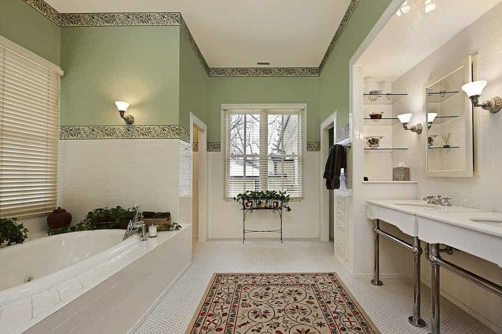 宁静的浴室，绿色的墙壁内衬绿叶壁纸。白色六边形地砖上铺着一条复古地毯，还有一个浴缸和一个双瓷洗手台。
