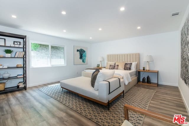 这间主卧室的特色是一张灰褐色的床，配上一张地毯上的白色沙发凳。白色天花板上的嵌壁式照明非常漂亮。