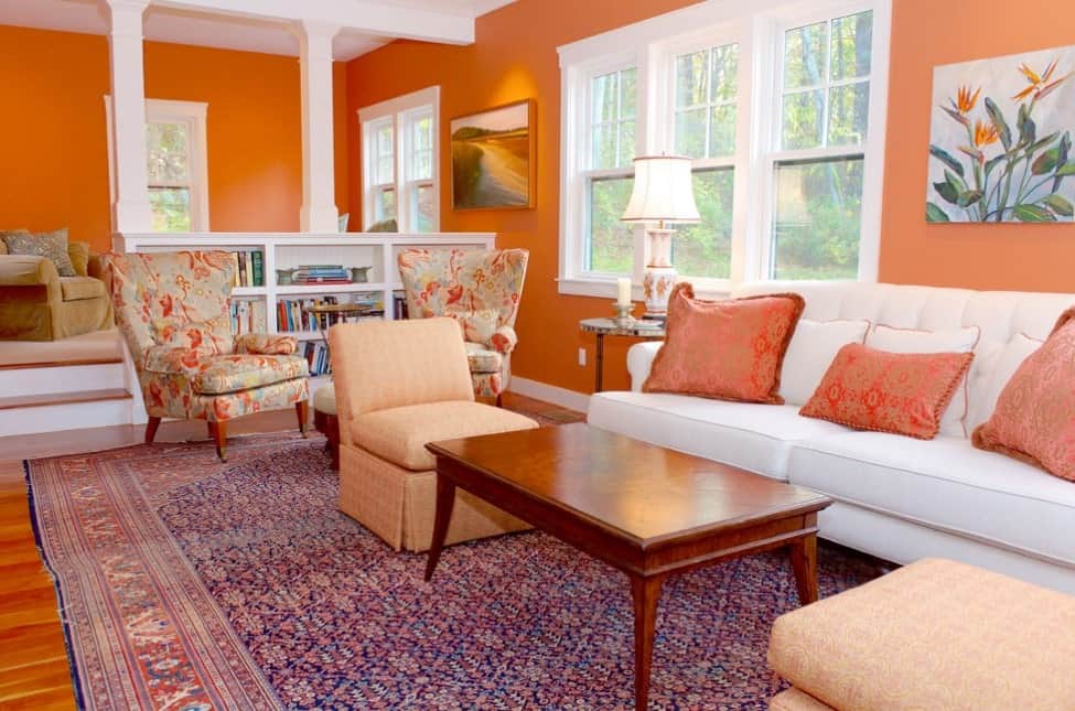 这个过渡的柿子橙色下沉客厅与白色的色调结合了切斯特菲尔德沙发的优雅，红色和白色抱枕，橙色和蓝色刺绣的高靠背椅，棕褐色和橙色座椅，古董东方地毯和灯，宁静的绘画，硬木地板，和白色漆窗。