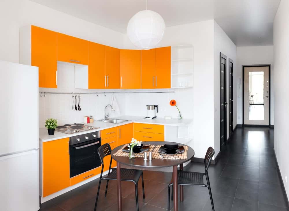 一个小的l型厨房，橙色的橱柜，一个简单的白色吊灯，和一个双人用餐桌，非常适合情侣。