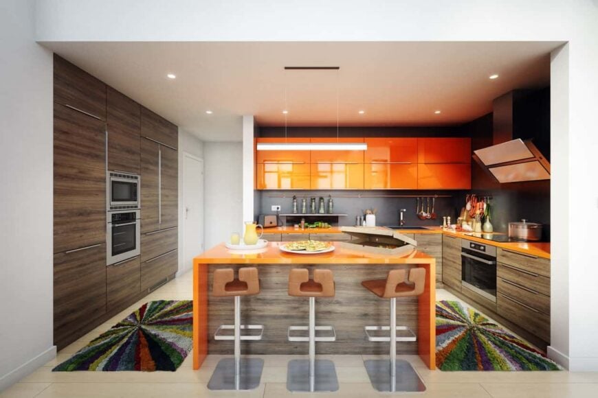 l型厨房配有早餐岛、酒吧式凳子、简单的直吊灯、闪亮的橙色橱柜和不锈钢电器。