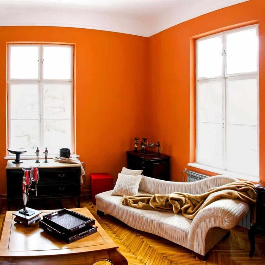 温暖的珊瑚橙色房间，配有舒适的躺椅，东方角柜，木桌和人字纹木地板。