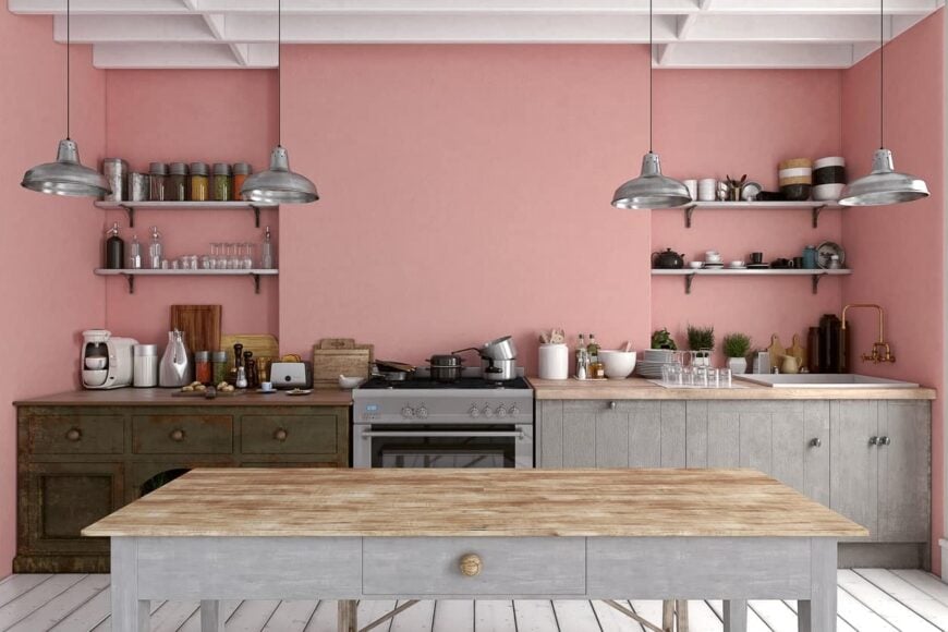 鲑鱼粉色的厨房，白色木梁天花板和木板地板。厨房柜台对面有一张木桌，上面挂着银色的吊灯。
