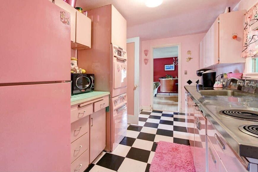 厨房设有浅粉色橱柜和不锈钢柜台，配有水槽和灶台。它的地板是格子的，上面有一块粉红色的蓬松的厨房地毯。