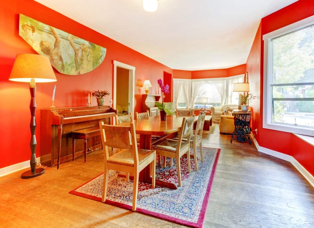 红色的餐厅里有一套可供六人用餐的木制餐具，上面铺着带边框的地毯，一盏传统的落地灯，一架钢琴在一幅古董墙艺术品下。