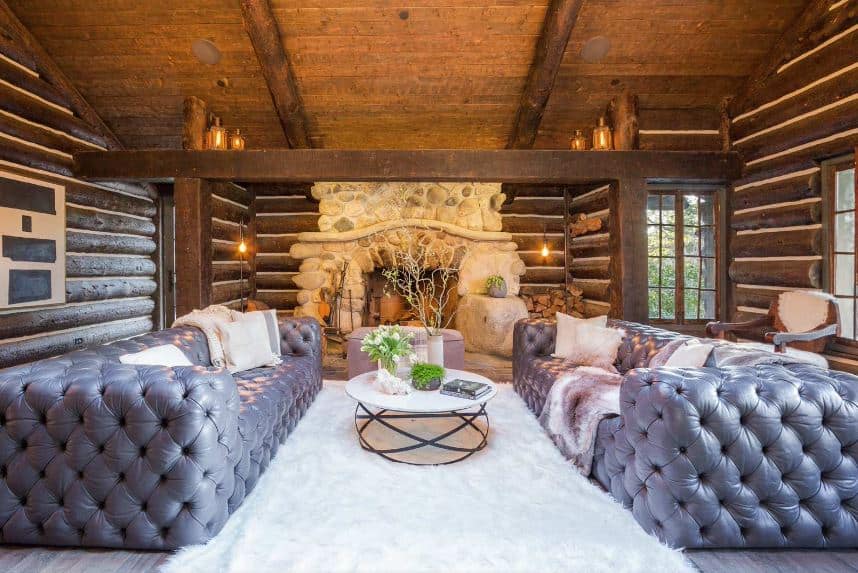 这个乡村风格的客厅的亮点是一对灰色簇绒沙发，两侧是白色圆形咖啡桌，上面是白色毛茸茸的小地毯。这个设置在木棚天花板上很突出，暴露的木梁与墙壁的原木梁相匹配。