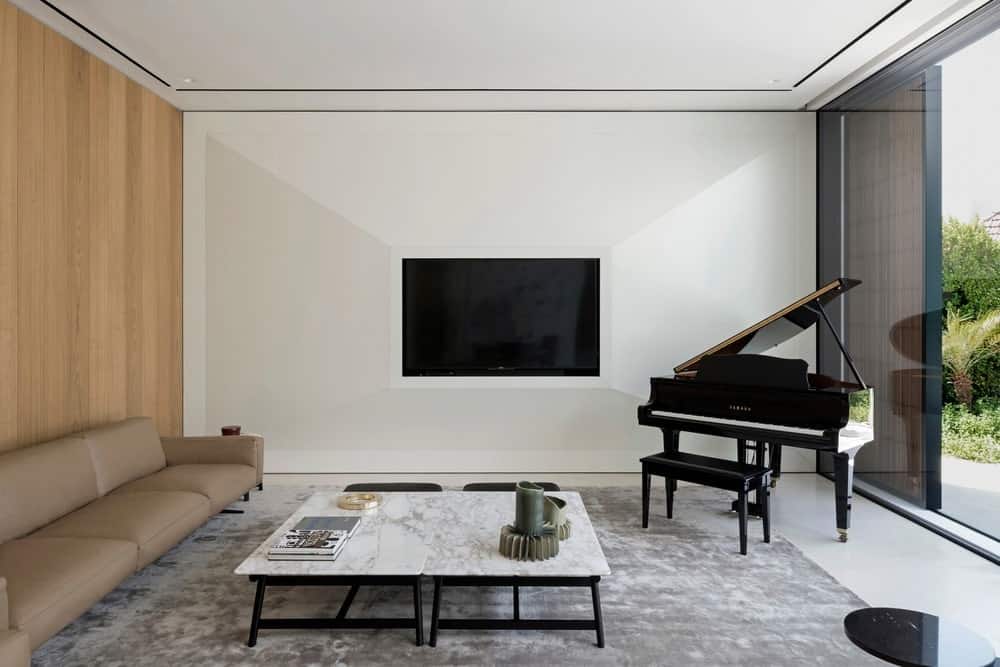 现代化的客厅里有一张米黄色组合沙发，靠在镶木板的墙上，还有大理石的咖啡桌，放在白色瓷砖地板上的灰色地毯上。它包括一台壁挂式电视和全景窗旁的小型三角钢琴，为室内带来充足的自然光线。