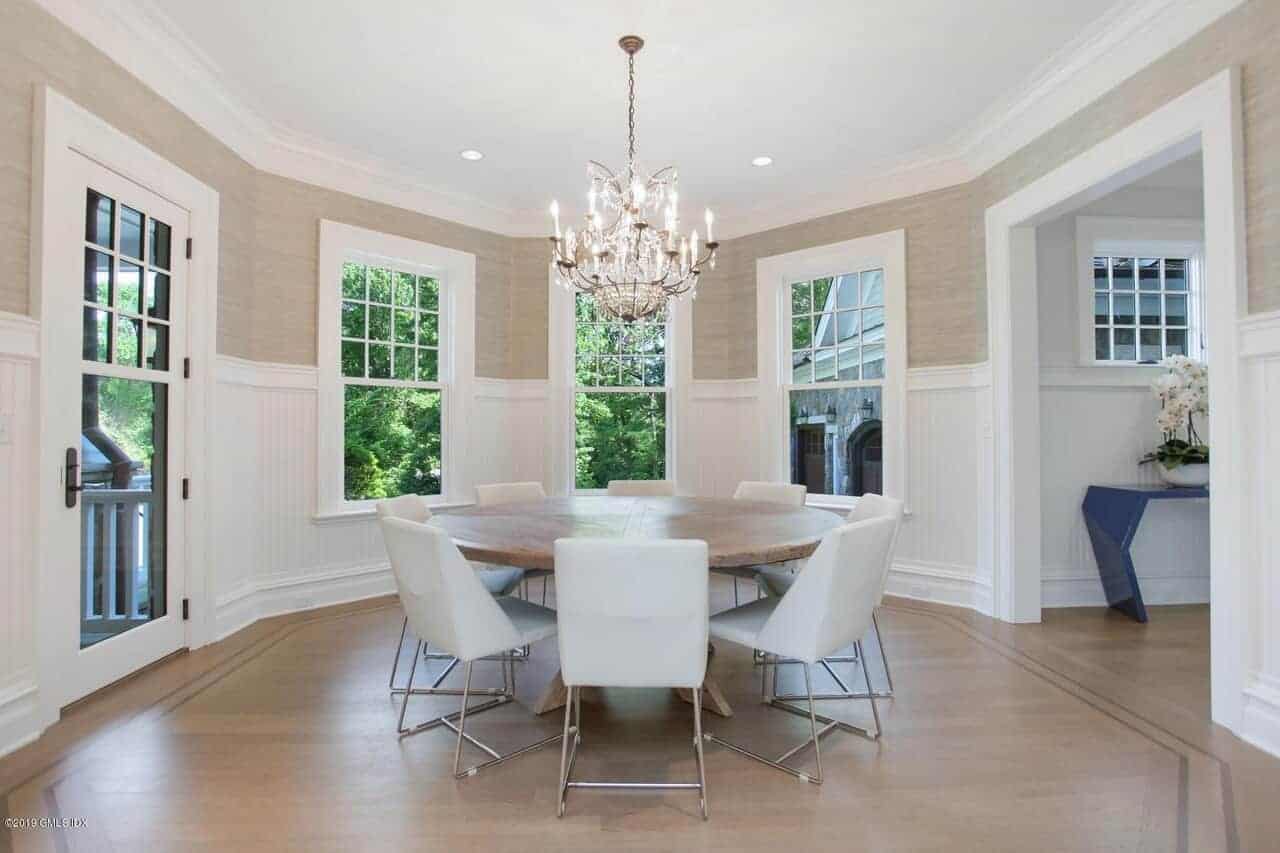 圆形木制餐桌周围环绕着现代的白色椅子，它们在硬木地板的映衬下显得格外突出，但与白色的护壁板相得益彰，优雅的枝形吊灯照亮了墙壁。