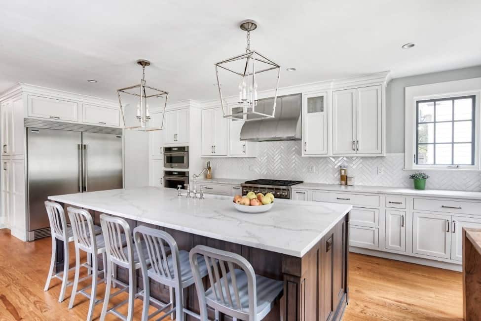 一个干净的白色厨房，有巨大的不锈钢冰箱，早餐岛，大理石台面，硬木地板，和吊灯。