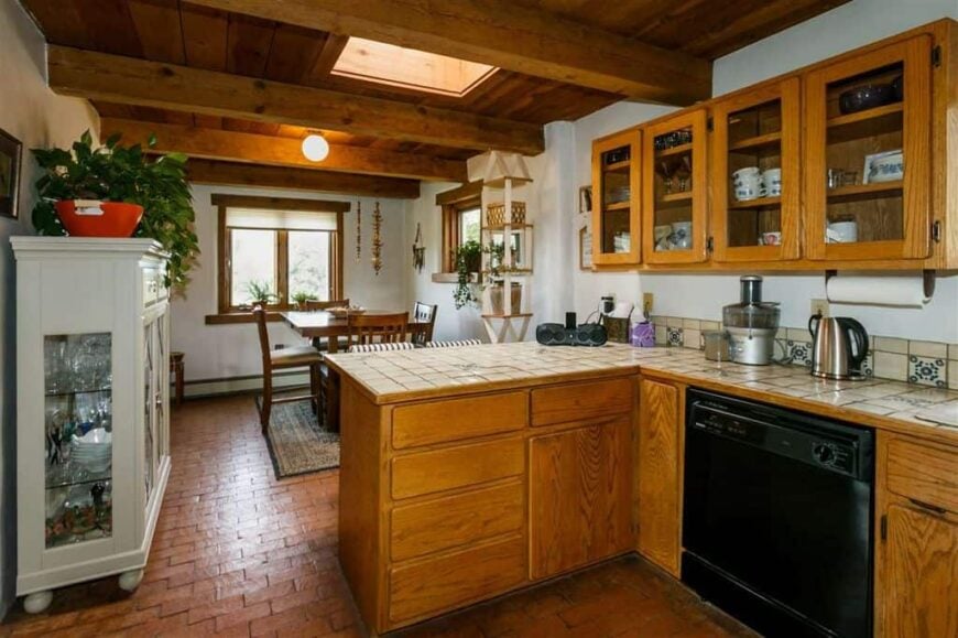 经典的l形厨房，砖地板和木制橱柜。它包括上部前部的玻璃柜和瓷砖台面。